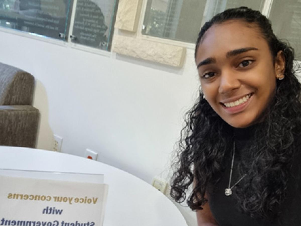 Analee Maharaj wears a black blouse and necklace and takes a selfie while sitting at a white table.